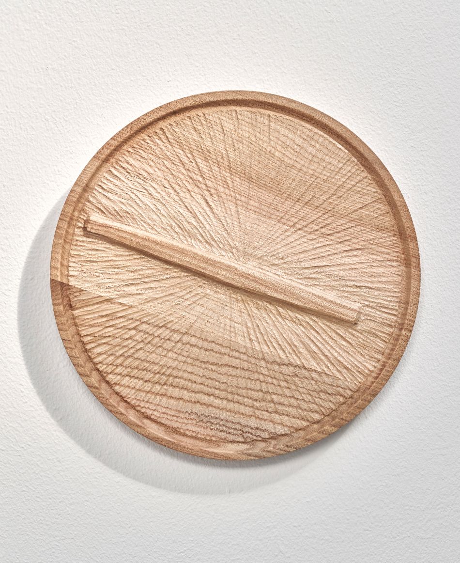 Image of Sculpture 'Now' made by Teresa Hunyadi. A wall-hung piece of a round relief carved piece reminiscent of a clock. The hands of the clock are appearing in one line.