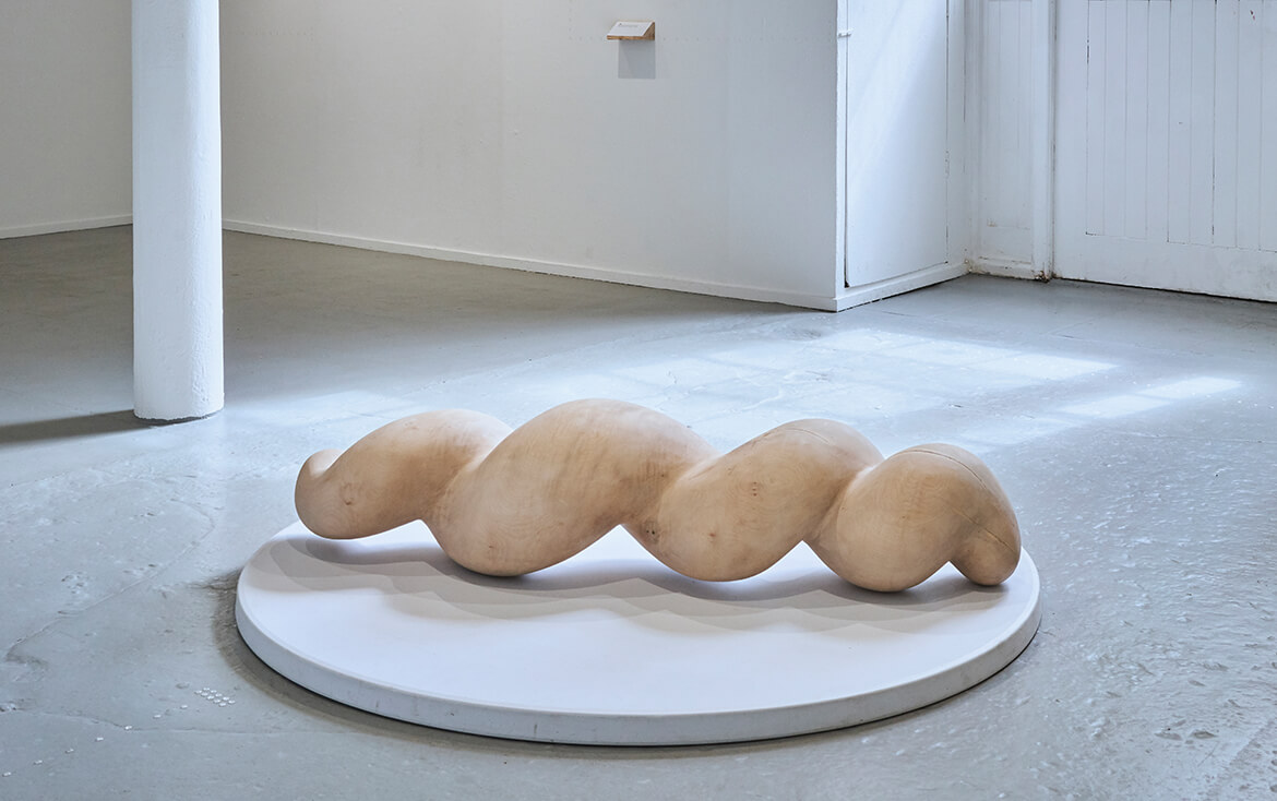 Big wood sculpture that is laying on a white platform. Its twisted shape makes it possible to be rolled from one side to the other. Teresa Hunyadi