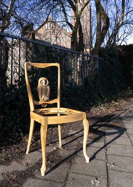 Wood sculpture of a bird (owl) and a chair placed outside.